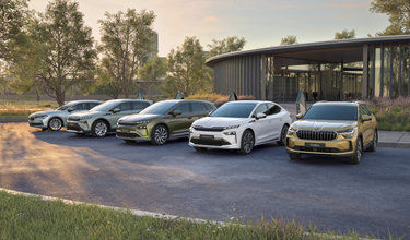 Four Škoda vehicles — including the Enyaq, Enyaq Coupé, and Kodiaq — displayed together on a forecourt outside a modern curved architectural building, surrounded by mature trees in a parkland setting at golden hour.